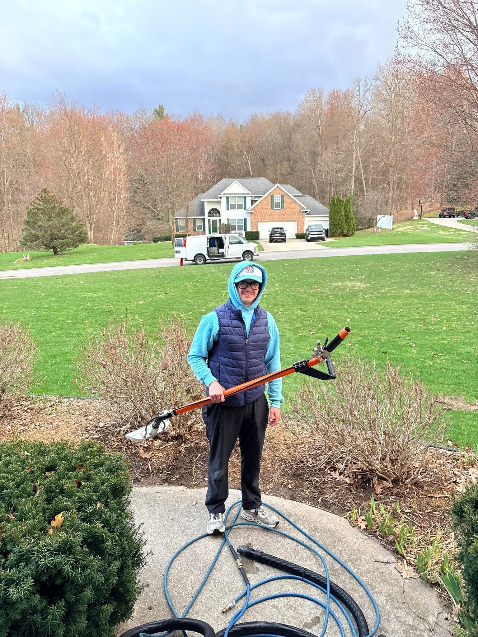 Person in blue jacket holding a long tool on a driveway, with a house and yard in the background
