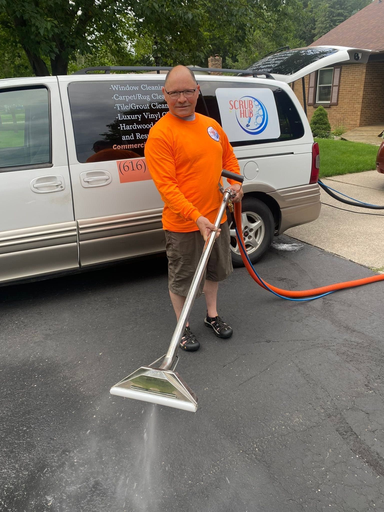 Person using a carpet-cleaning wand beside a van, cleaning wet pavement outside a house.