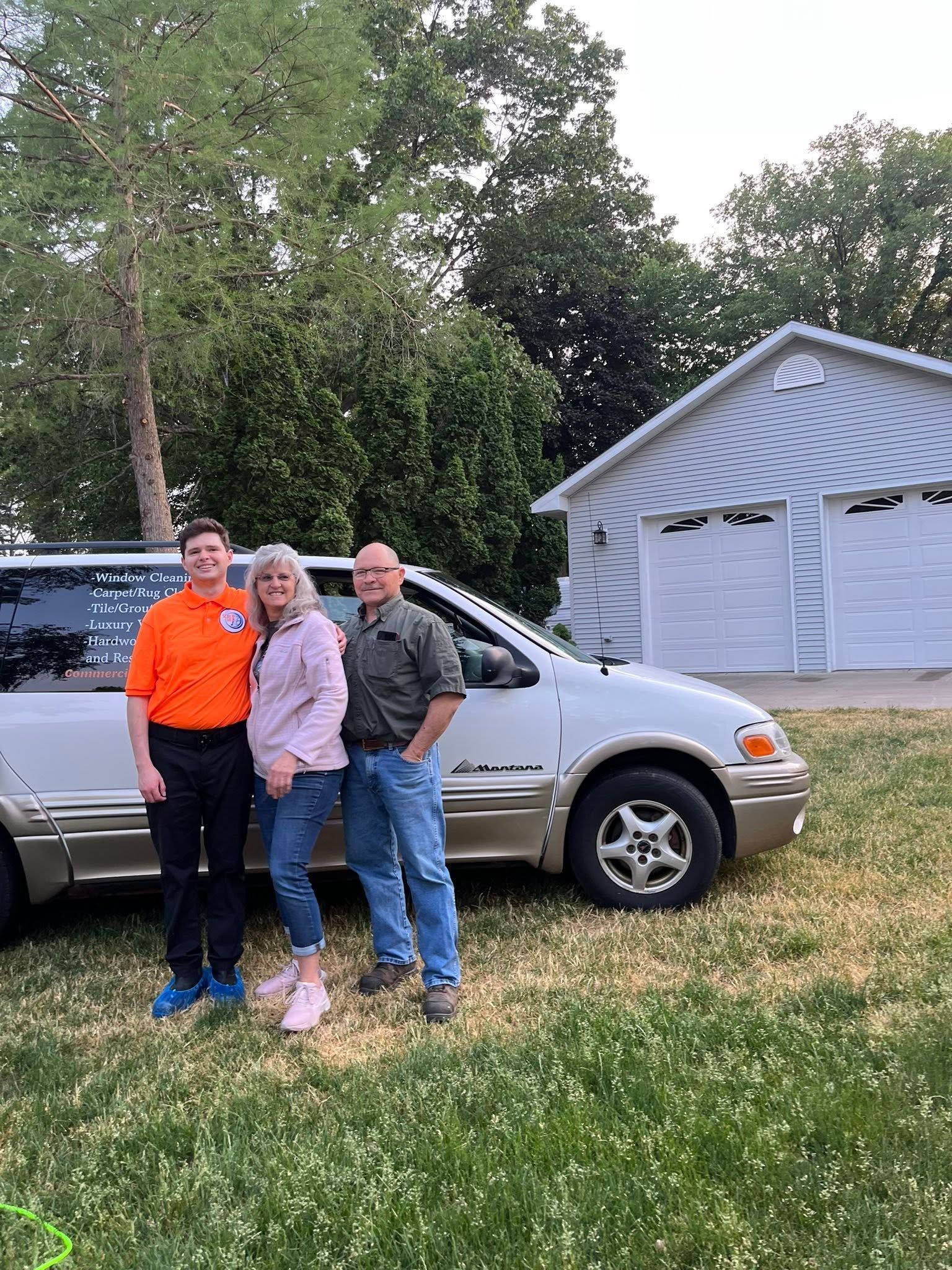 Three people standing in front of a silver SUV beside a white garage in a grassy yard.
