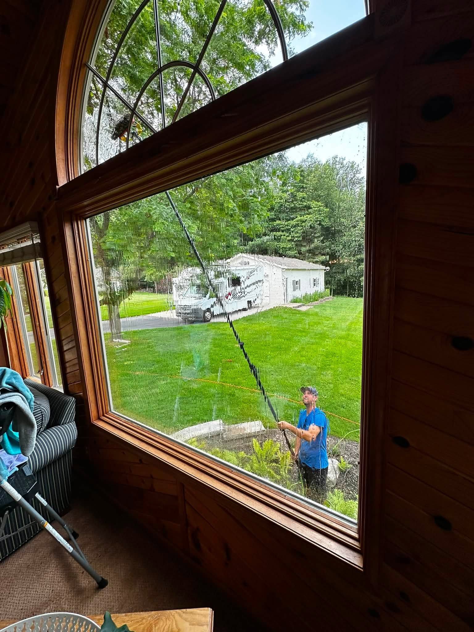 Man cutting branches in a green backyard, seen through a porch window with a chair inside.