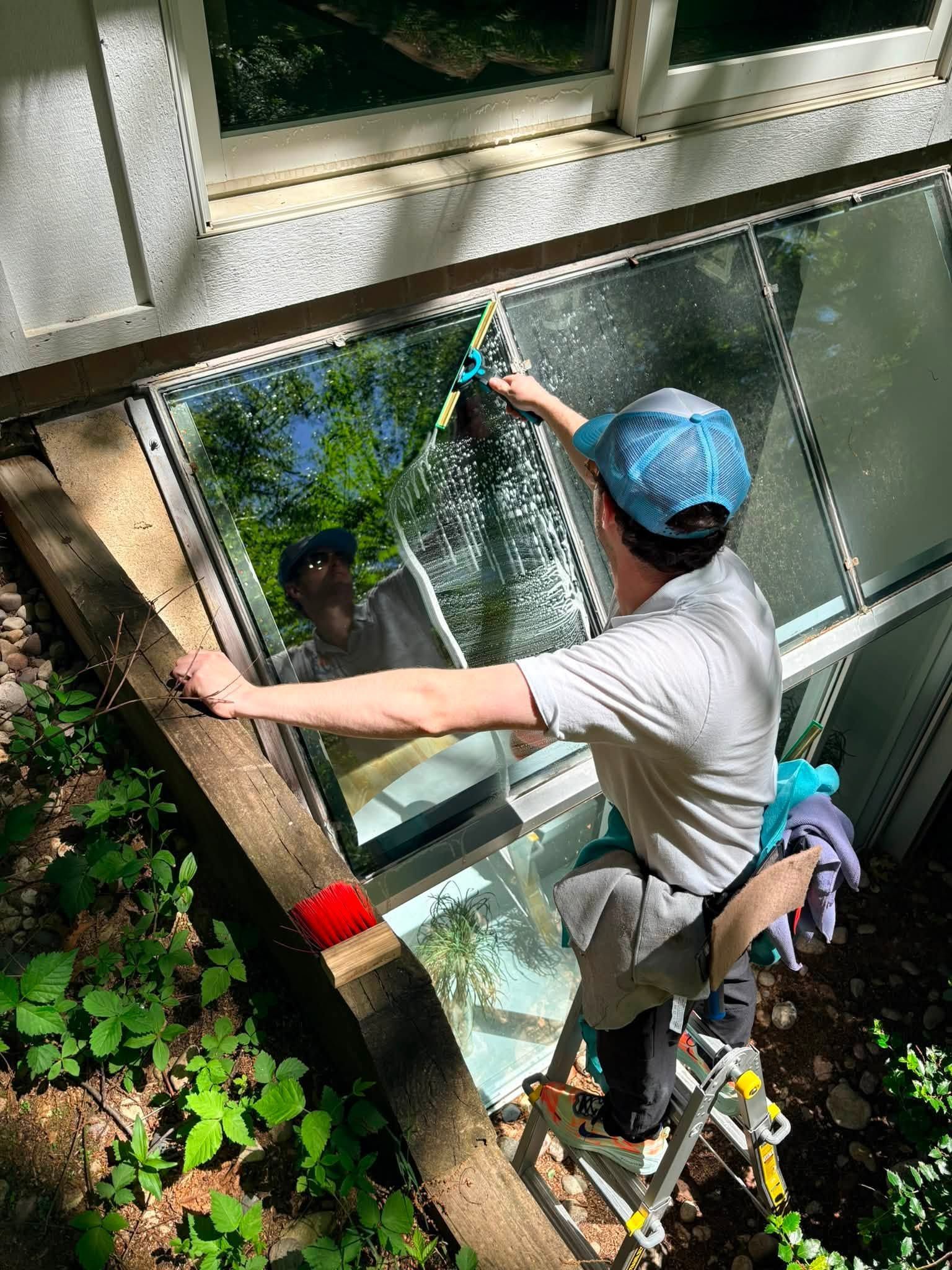 Worker in blue helmet cleaning an exterior window with a squeegee from a ladder
