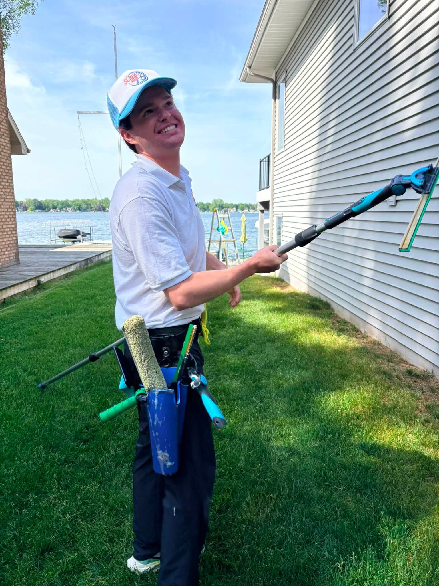 Person smiling outdoors while holding a pressure-washer wand beside a house, wearing a cap and work belt