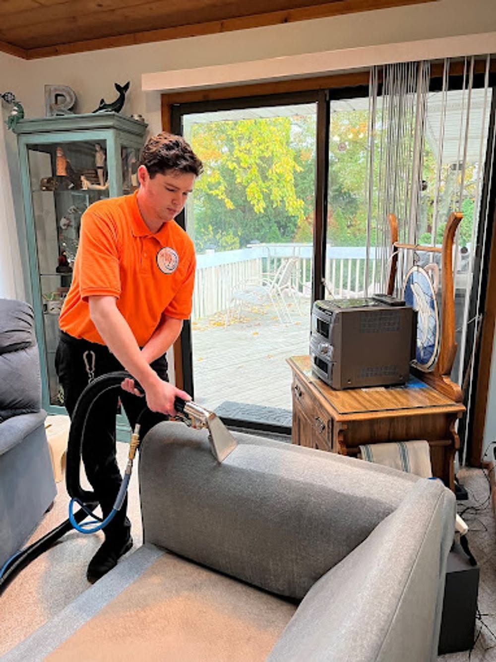 Person in orange shirt vacuuming a gray couch in a living room near a sliding glass door