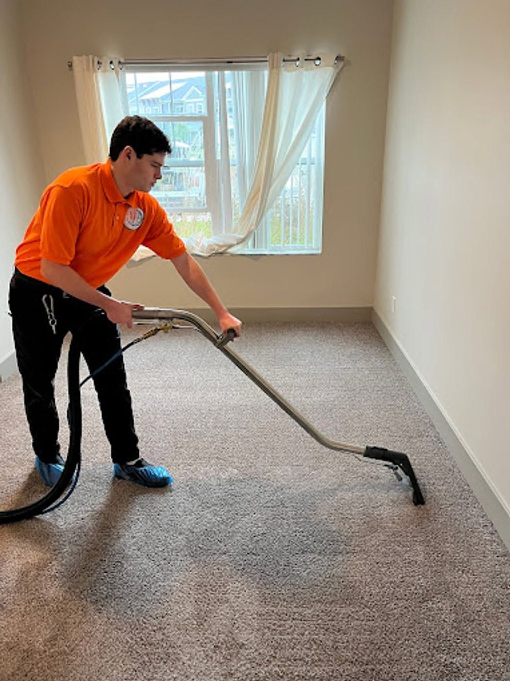 Worker vacuuming carpet in an empty room with a window and white curtains.
