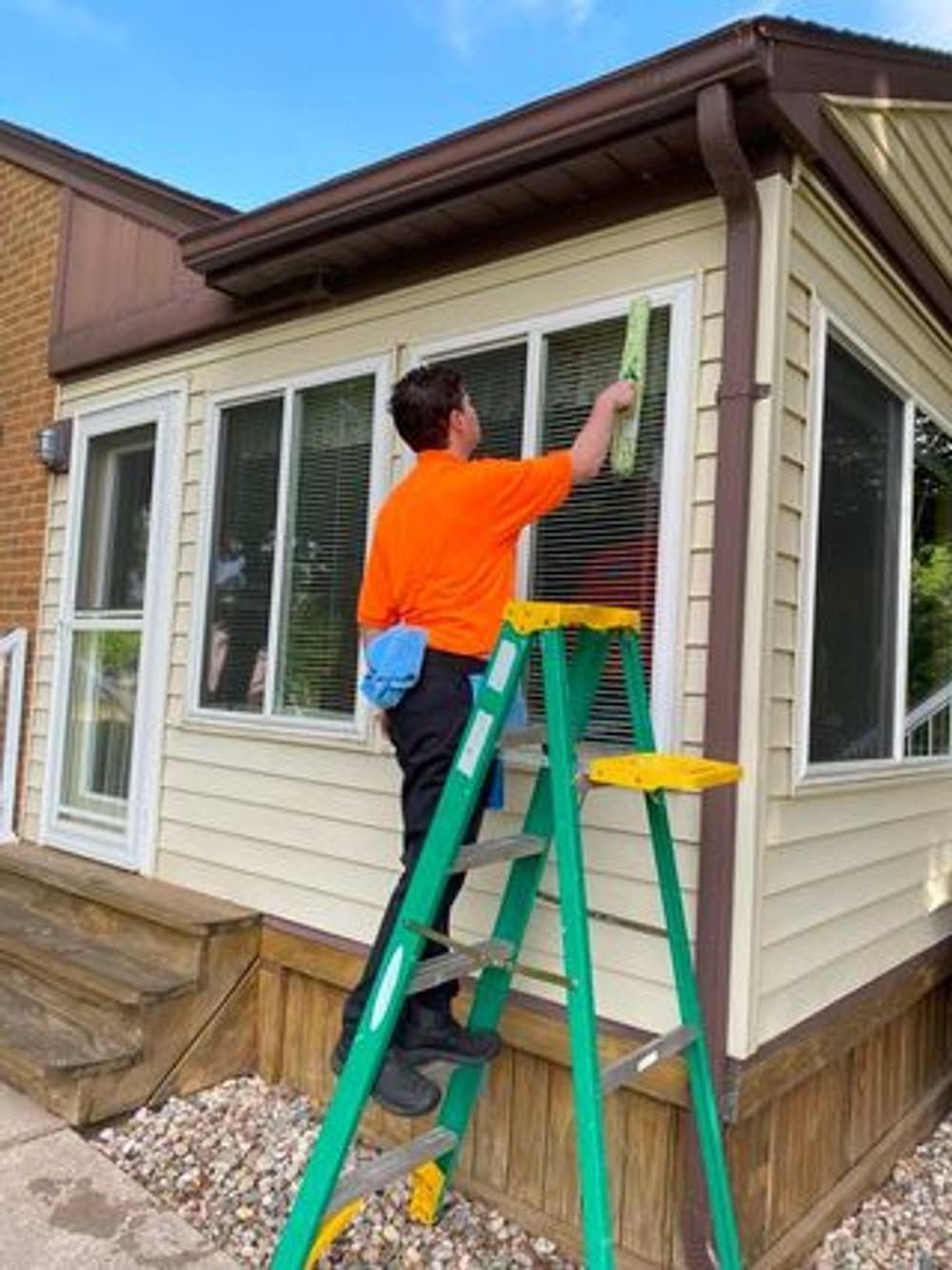 Person in orange shirt cleaning exterior window with a green ladder beside a house.