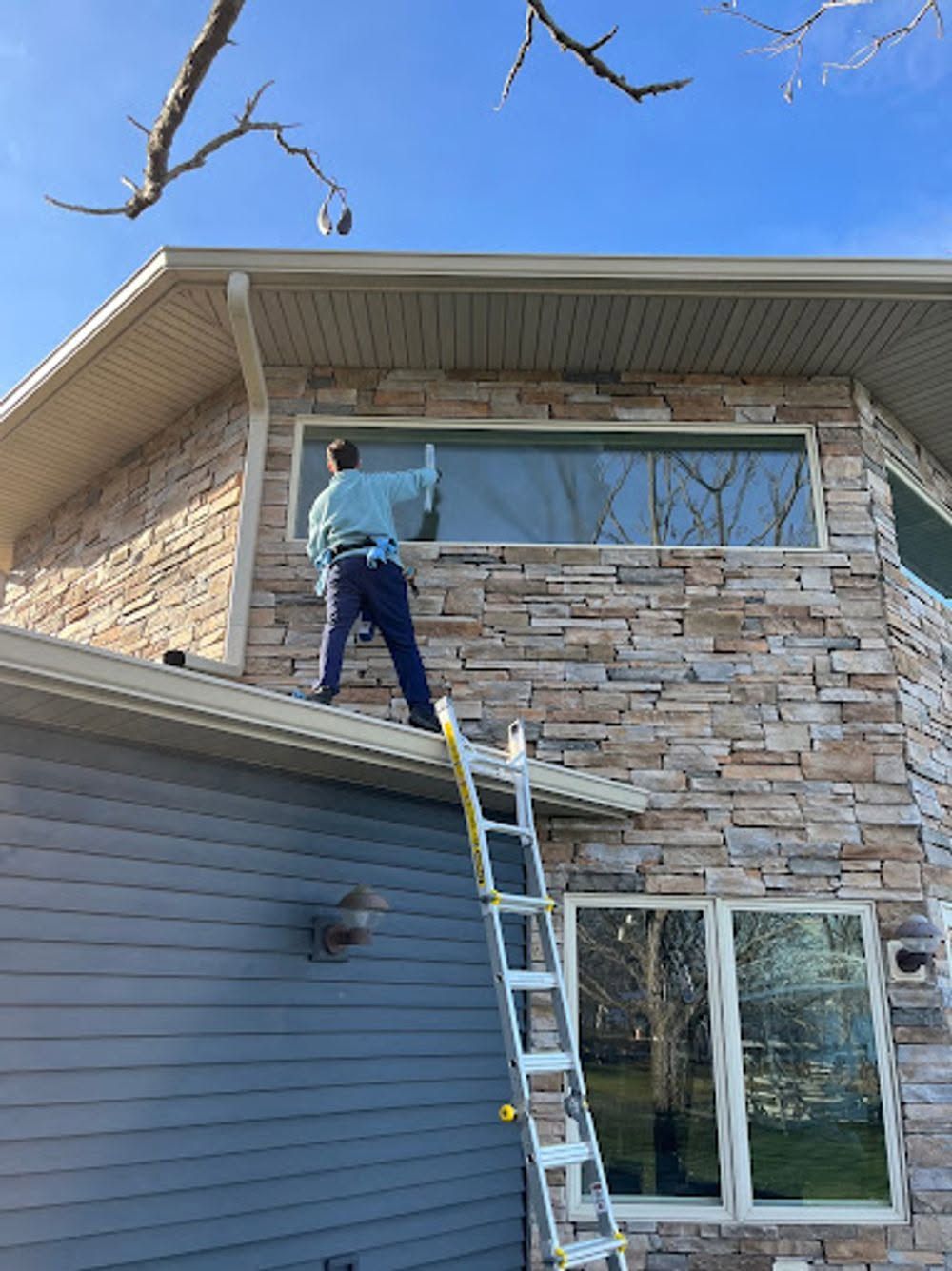 Worker on a ladder cleaning a large second-story window of a stone house