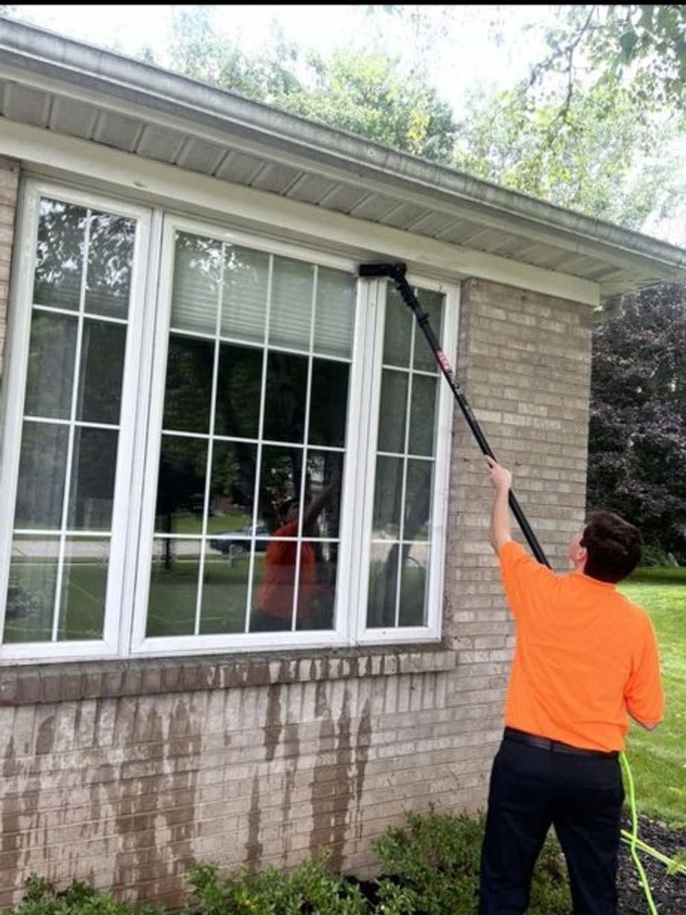 Person using a long-handled brush to clean the exterior windows and soffit of a brick house.