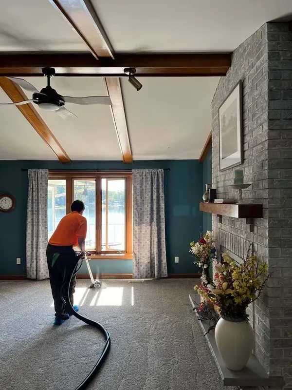Person vacuuming a bright living room with teal walls, wooden beams, and a stone fireplace.