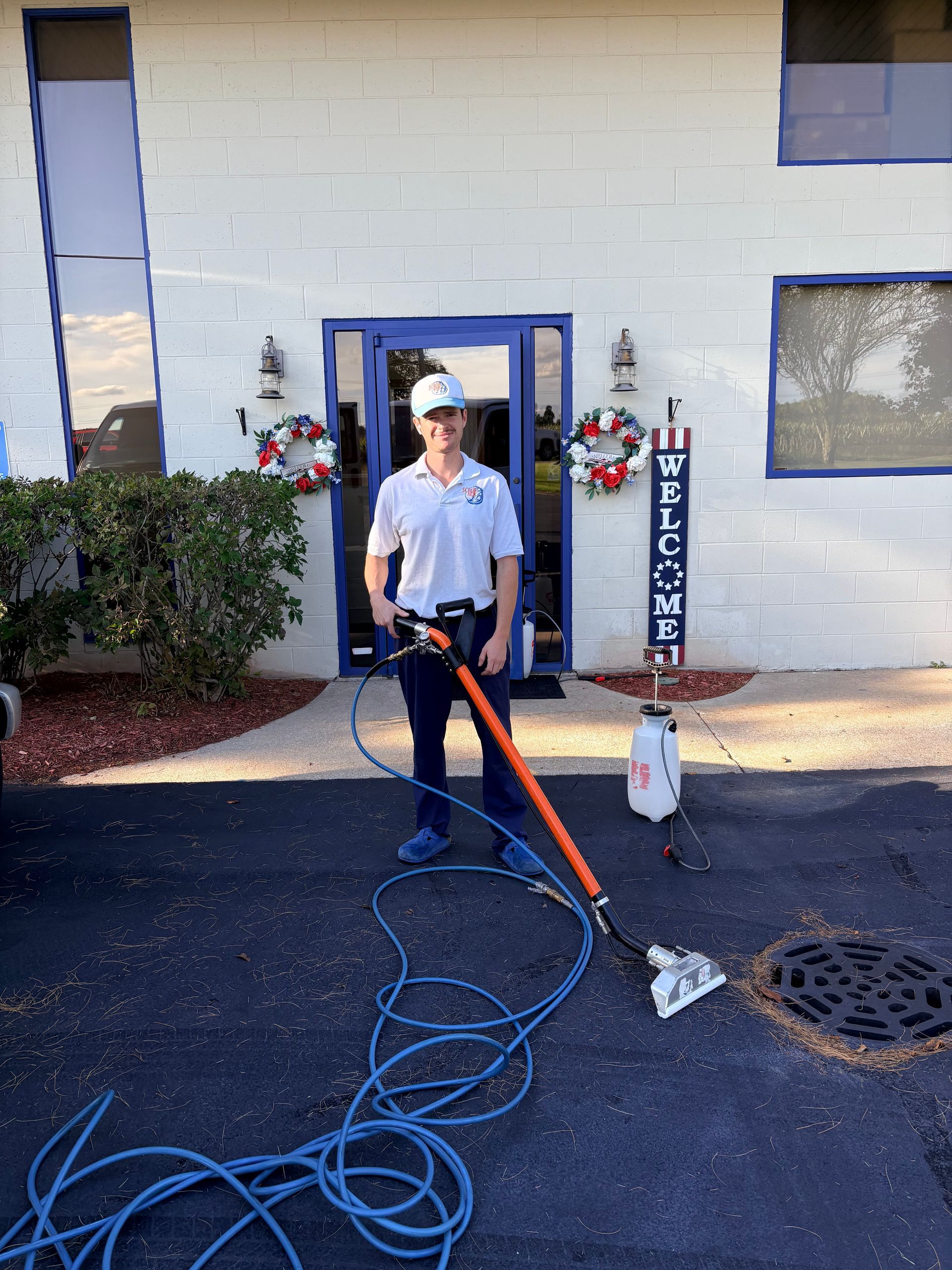 Worker using a power washer on a building exterior, with blue hose coiled on the ground.