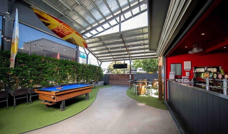 Covered outdoor patio with picnic tables, green turf, bar counter, and plants under a metal roof