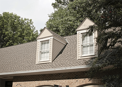 Brick house roof with gray shingles and two dormer windows surrounded by trees