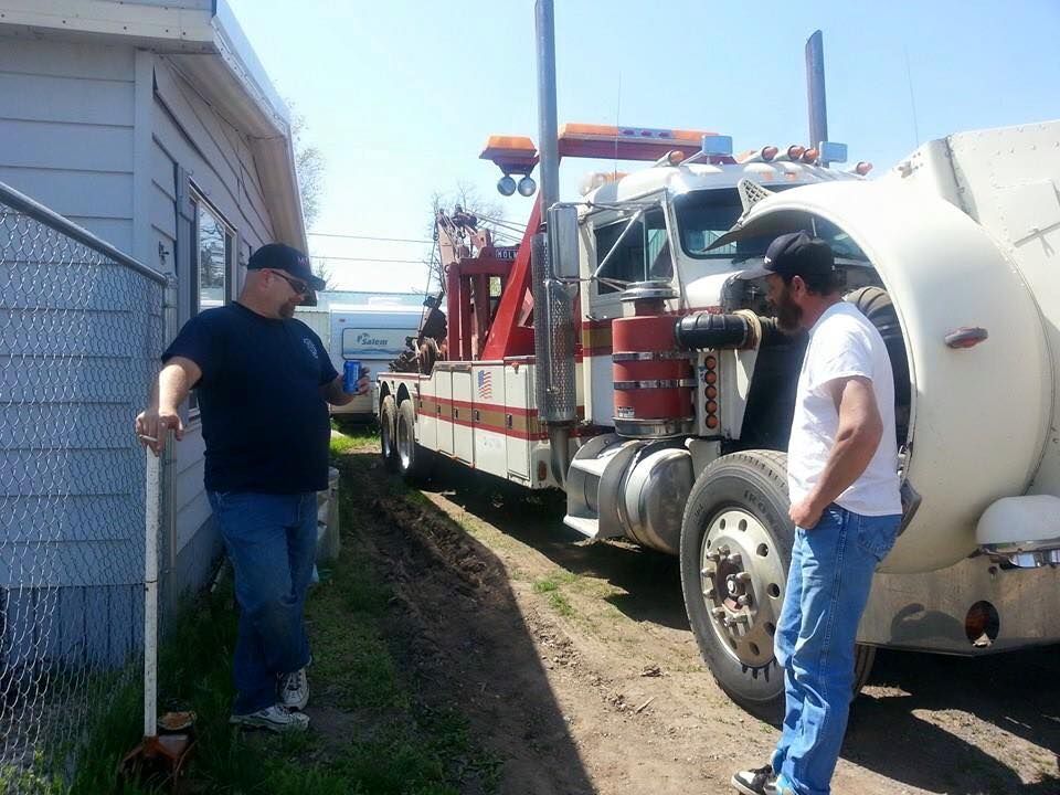 Two individuals stand outdoors talking next to a large white and red tow truck parked on a dirt path beside a building.