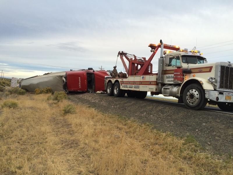 A heavy-duty tow truck is attached to a red semi-truck that has overturned on its side in a grassy field.