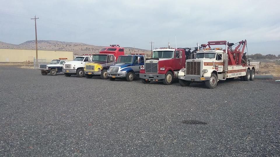 Six work trucks, including several tow trucks, parked in a line on a gravel lot under a cloudy sky.