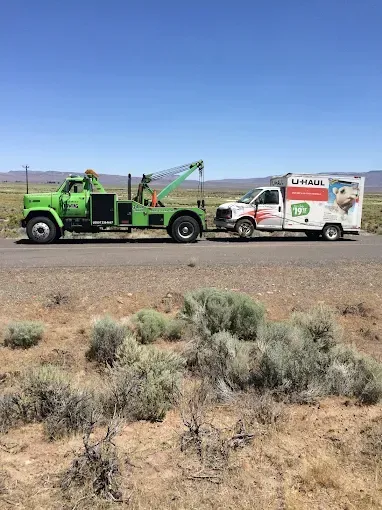 A bright green tow truck tows a white U-Haul moving truck along a desolate, sunlit desert road.