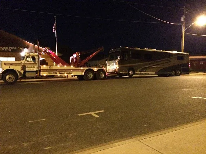 A tow truck at night pulling a large recreational vehicle along a dark street.