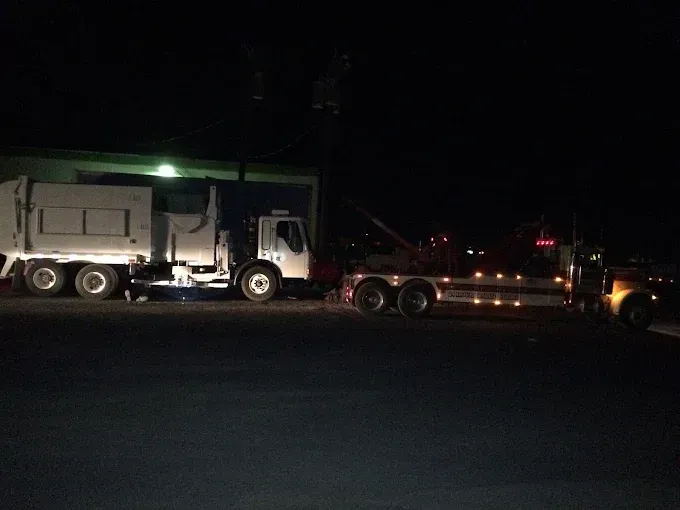 A white garbage truck being towed by a wrecker truck at night.