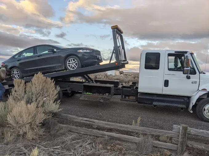 A dark gray sedan is being towed on a flatbed truck parked on a road near dry desert brush at sunset.