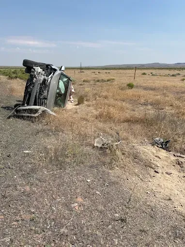 A silver vehicle lying on its side in dry, grassy terrain next to a dirt road under a clear sky.