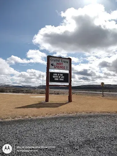 A sign for Crystal Crane Hot Springs stands on a dry, grassy field under a bright, cloudy sky.