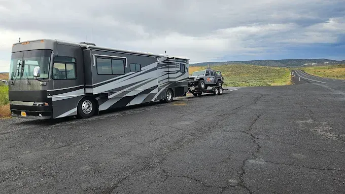 A gray and black motorhome towing a pickup truck parked on the side of a paved road in a dry, open landscape.