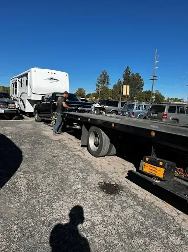 A person prepares to load a dark pickup truck, towing a white camper, onto a flatbed tow truck in a sunny, paved lot.
