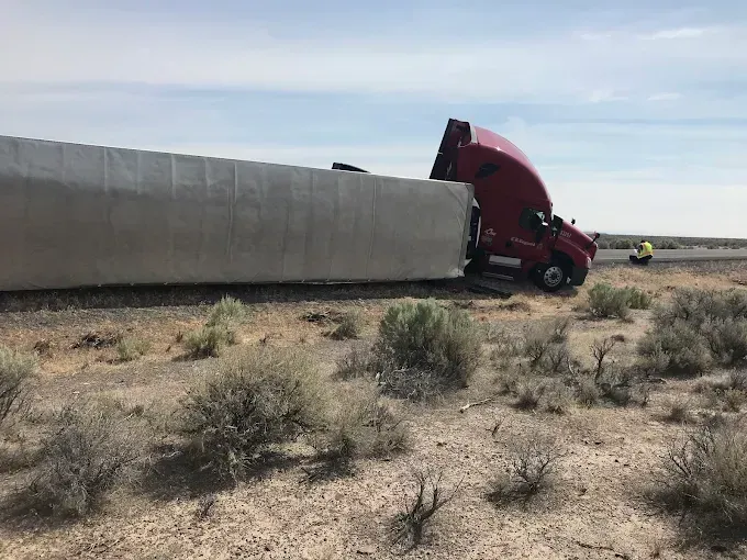 A red semi-truck lies on its side in the dirt off the shoulder of a highway, with a person standing nearby.