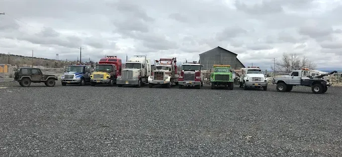 A row of diverse semi-trucks, heavy-duty vehicles, and a small jeep parked on a gravel lot under a cloudy sky.