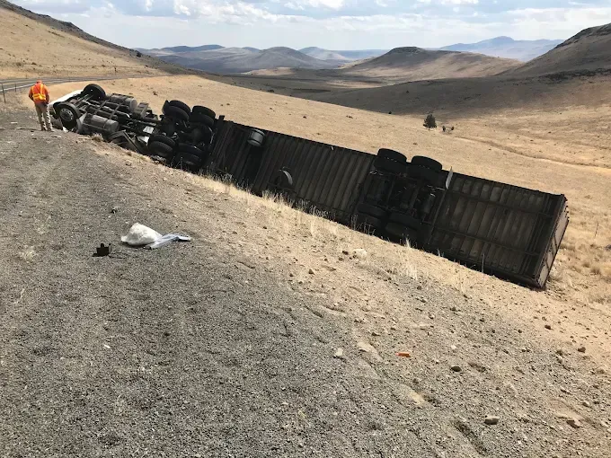 A semi-truck lies overturned on its side down a steep, dry embankment with a person in an orange vest standing nearby.