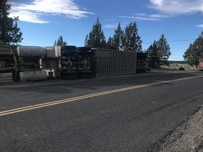 A semi-truck lies overturned on its side, blocking a two-lane paved road near a treeline under a bright blue sky.
