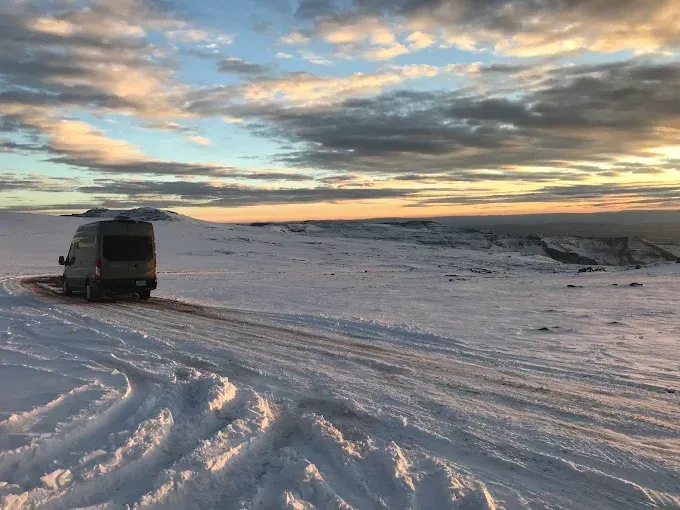 A van parked on a snowy road during a golden sunset, with vast, snow-covered landscape stretching into the distance.