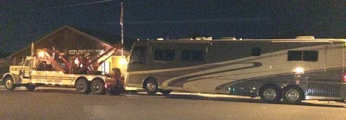 A tow truck pulling a large, tan-and-brown motorhome parked in front of a building at night.