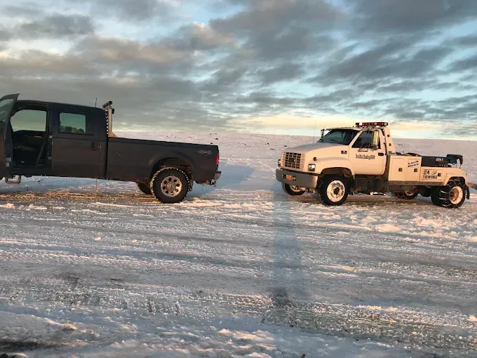 A black pickup truck parked next to a white tow truck on a snowy, open field under a cloudy sky.