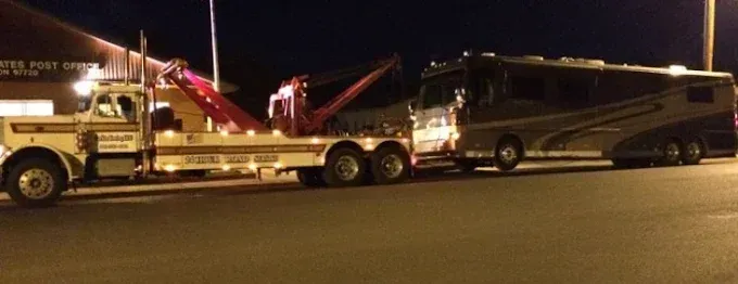 A white tow truck is towing a large, dark-colored recreational vehicle at night on an asphalt road.