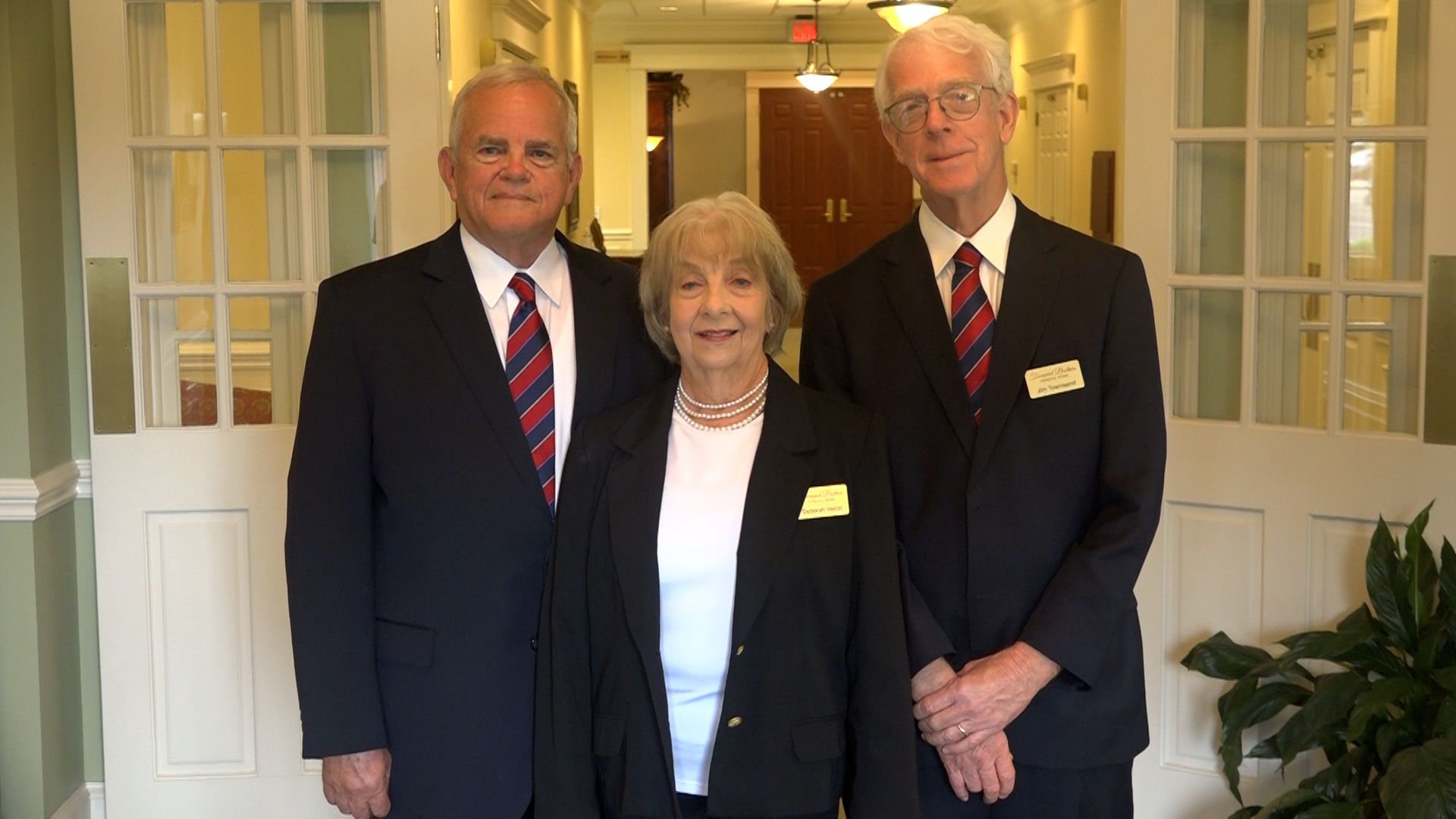 Three people in suits posing in a hallway. Two men flank a woman, all wearing dark jackets and red ties.
