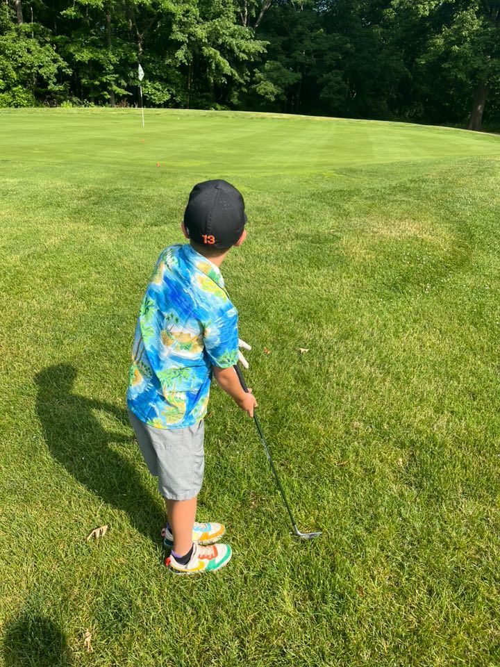 A golfer in a blue patterned shirt stands on a green grass course, holding a golf club and facing a distant flag.