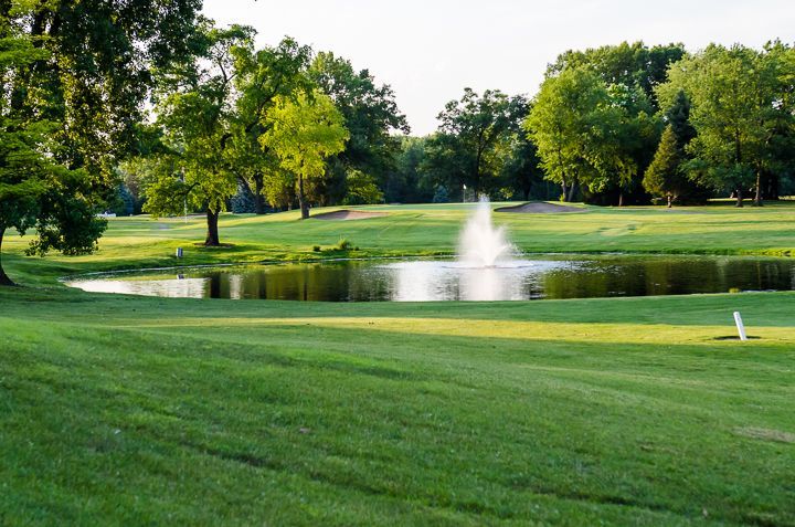 A golf course green with a small pond and fountain in the center, surrounded by lush green trees under a bright sky.