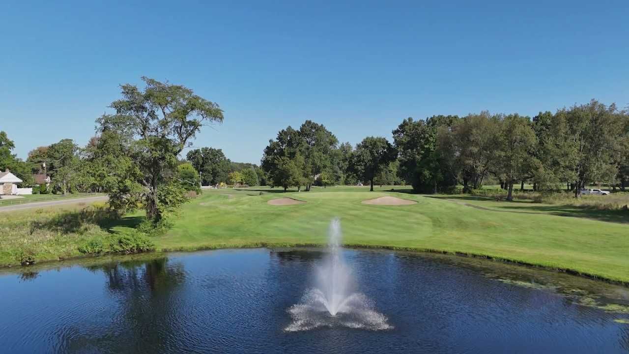 A water fountain sprays in the center of a calm pond, with a grassy golf course and trees visible in the background.