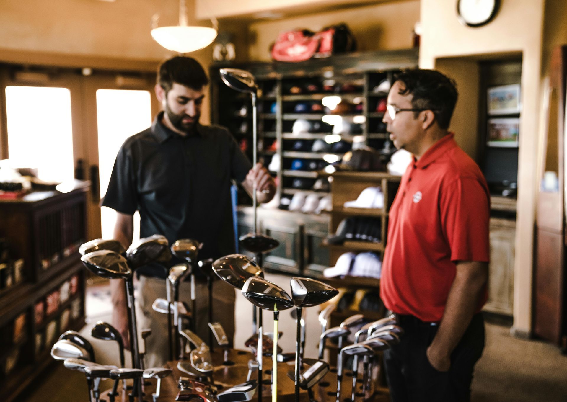 Two people discuss a golf club in a well-lit pro shop filled with shelves of equipment and apparel.