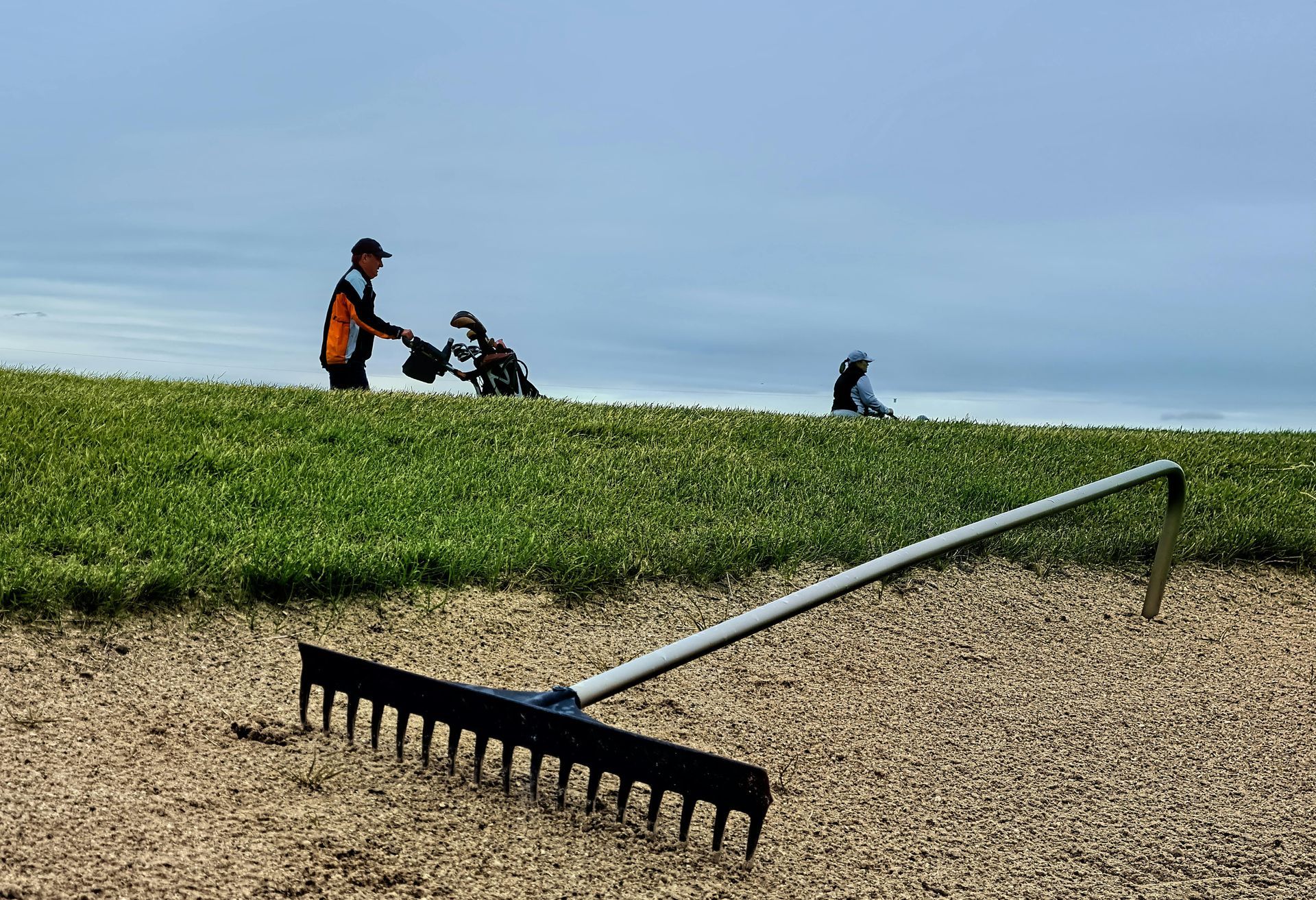 A sand rake lies in the foreground of a bunker, with golfers visible on the grassy hill in the distance.