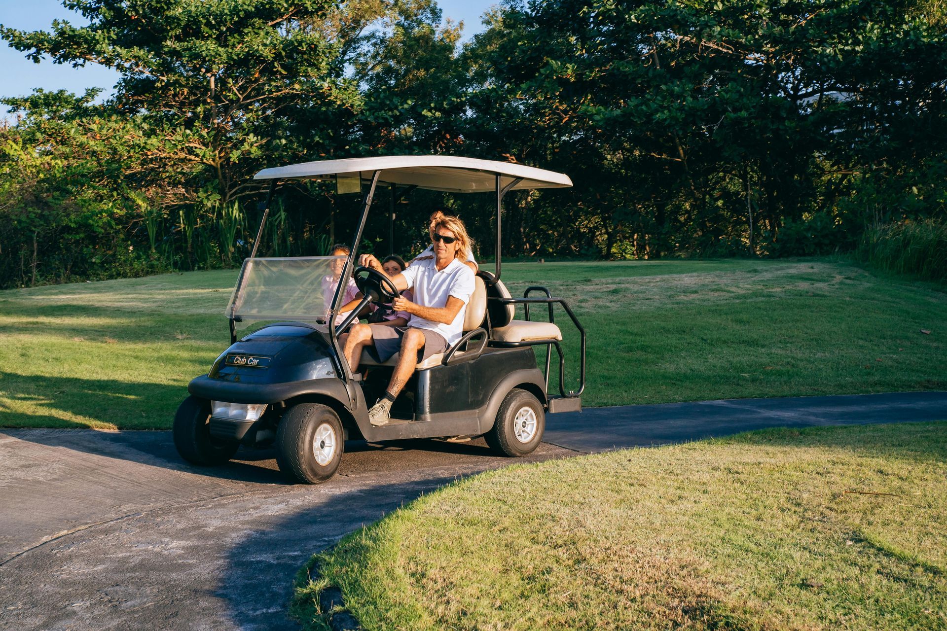 A person wearing sunglasses drives a black golf cart along a paved path bordered by green grass and trees.