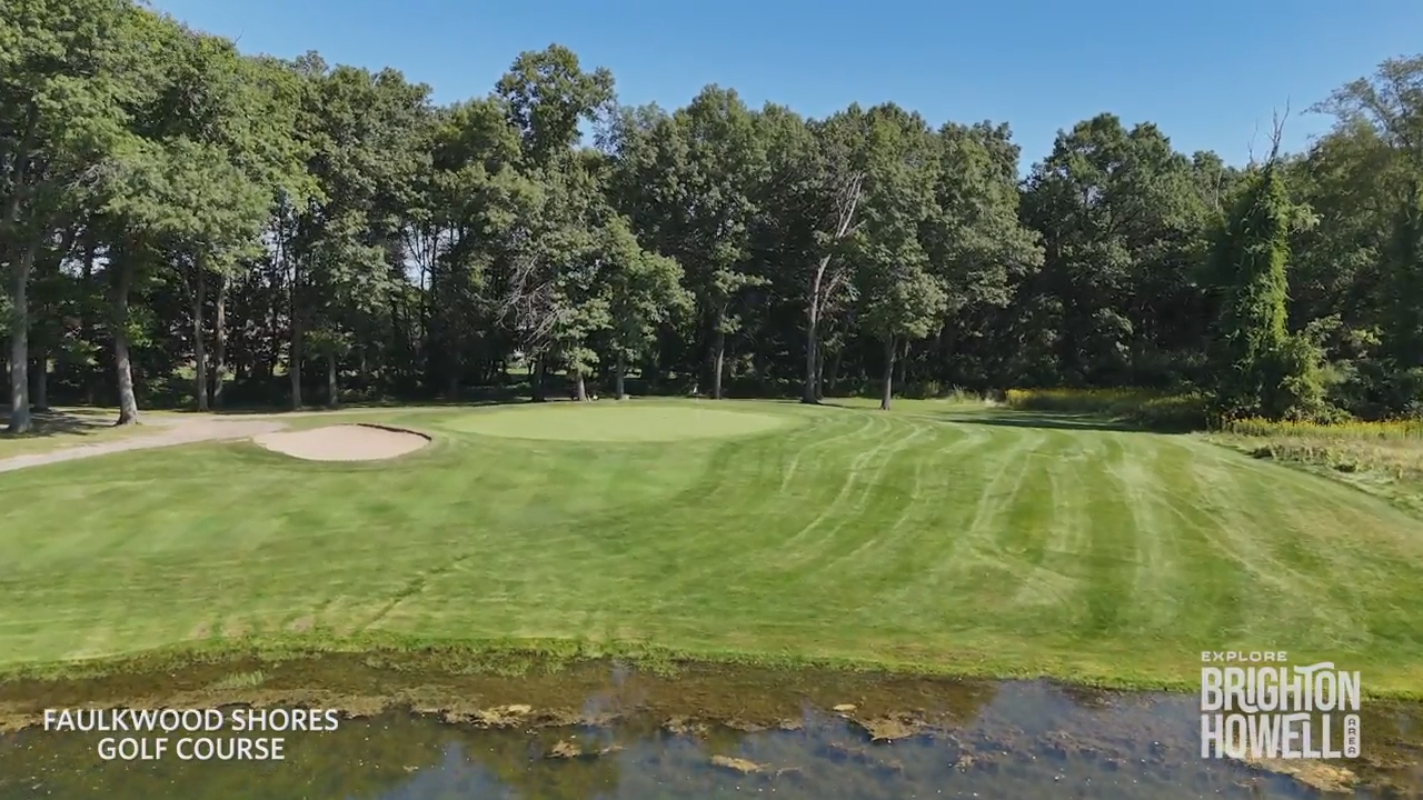 A green golf course with a pond in the foreground, sand bunker, and dense trees in the background under a blue sky.