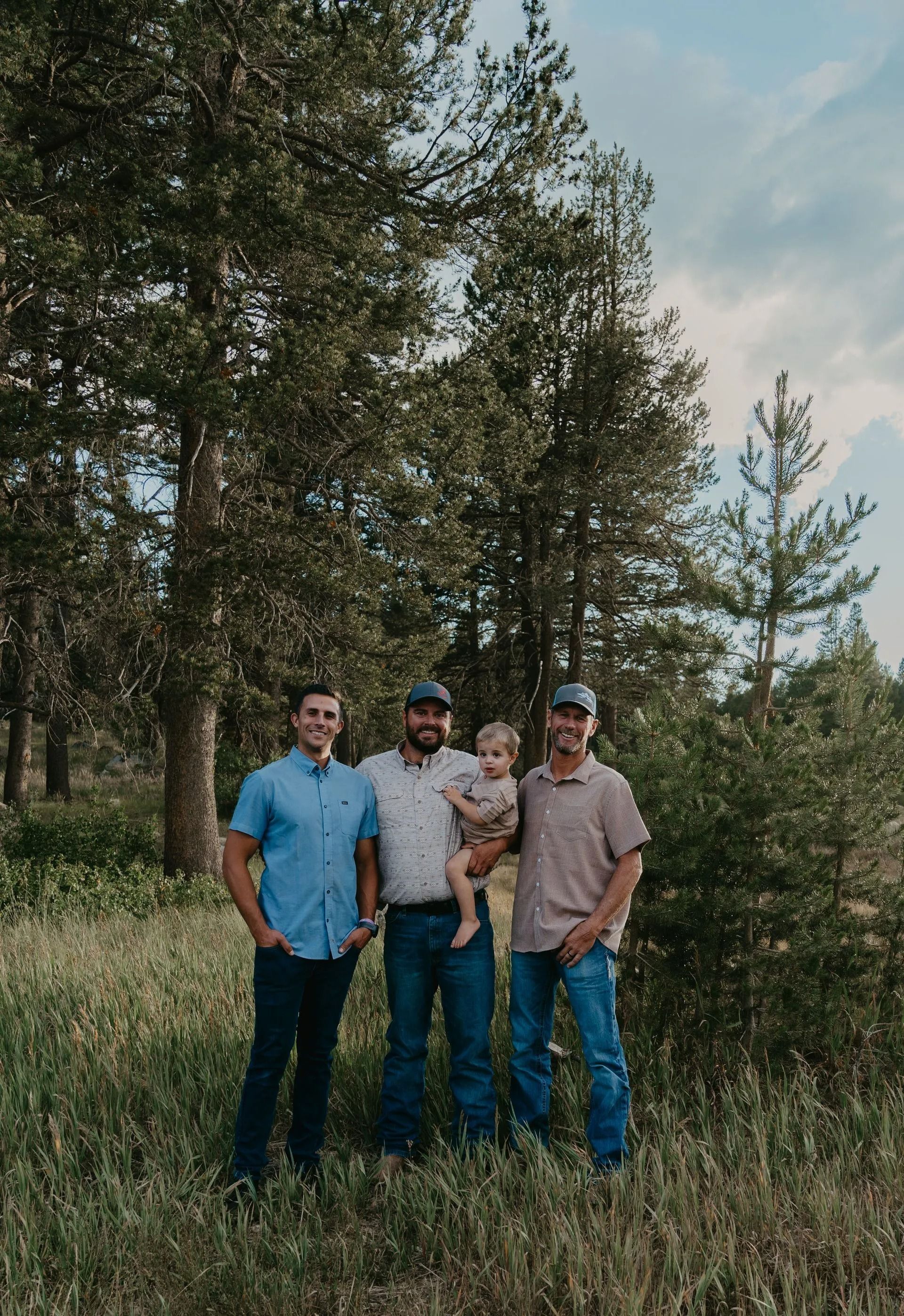 Four people standing in a grassy area with trees. One person holds a child. They wear casual clothing.