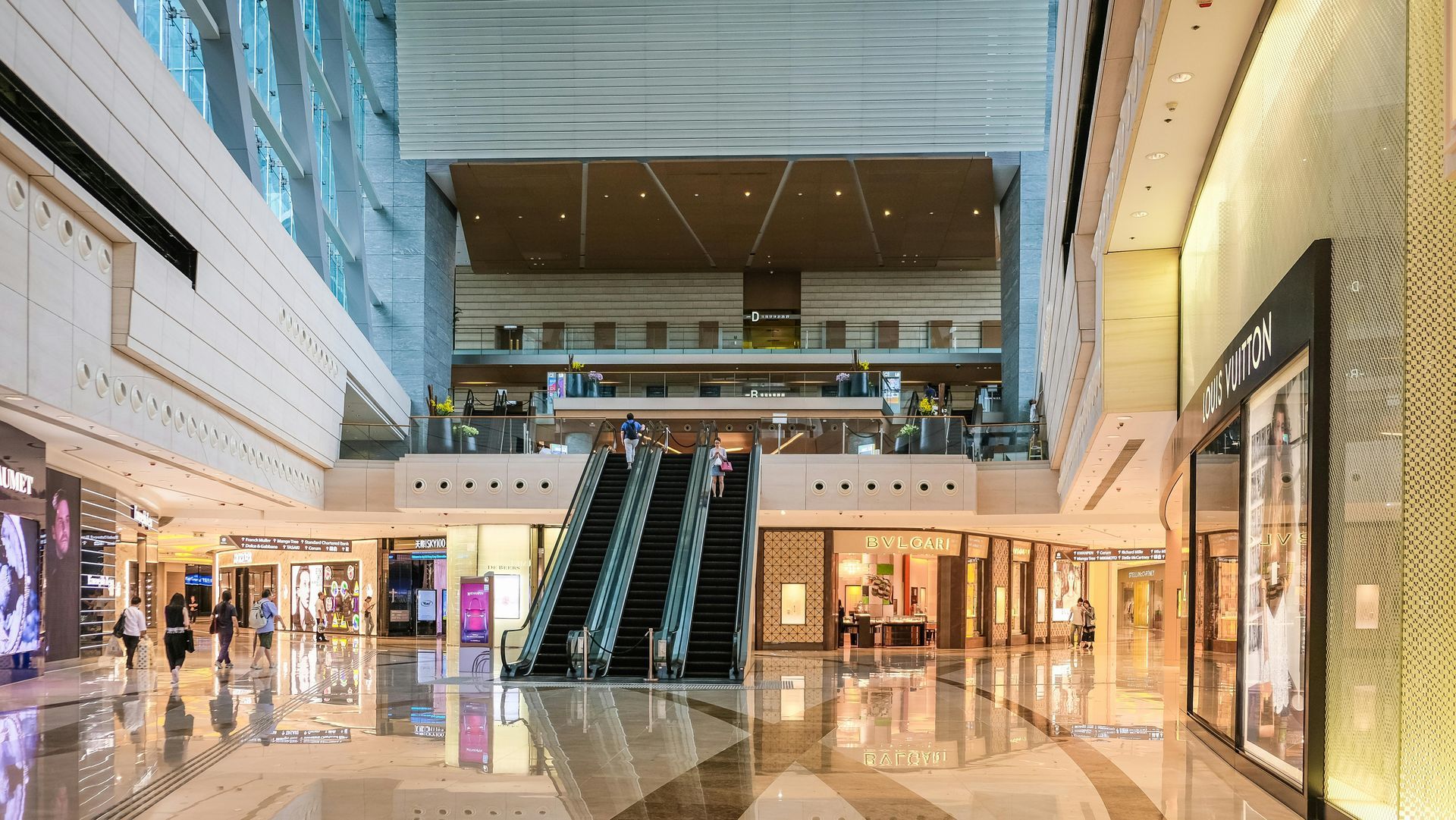 Interior of a modern shopping mall with escalators, retail shops, and people walking around.