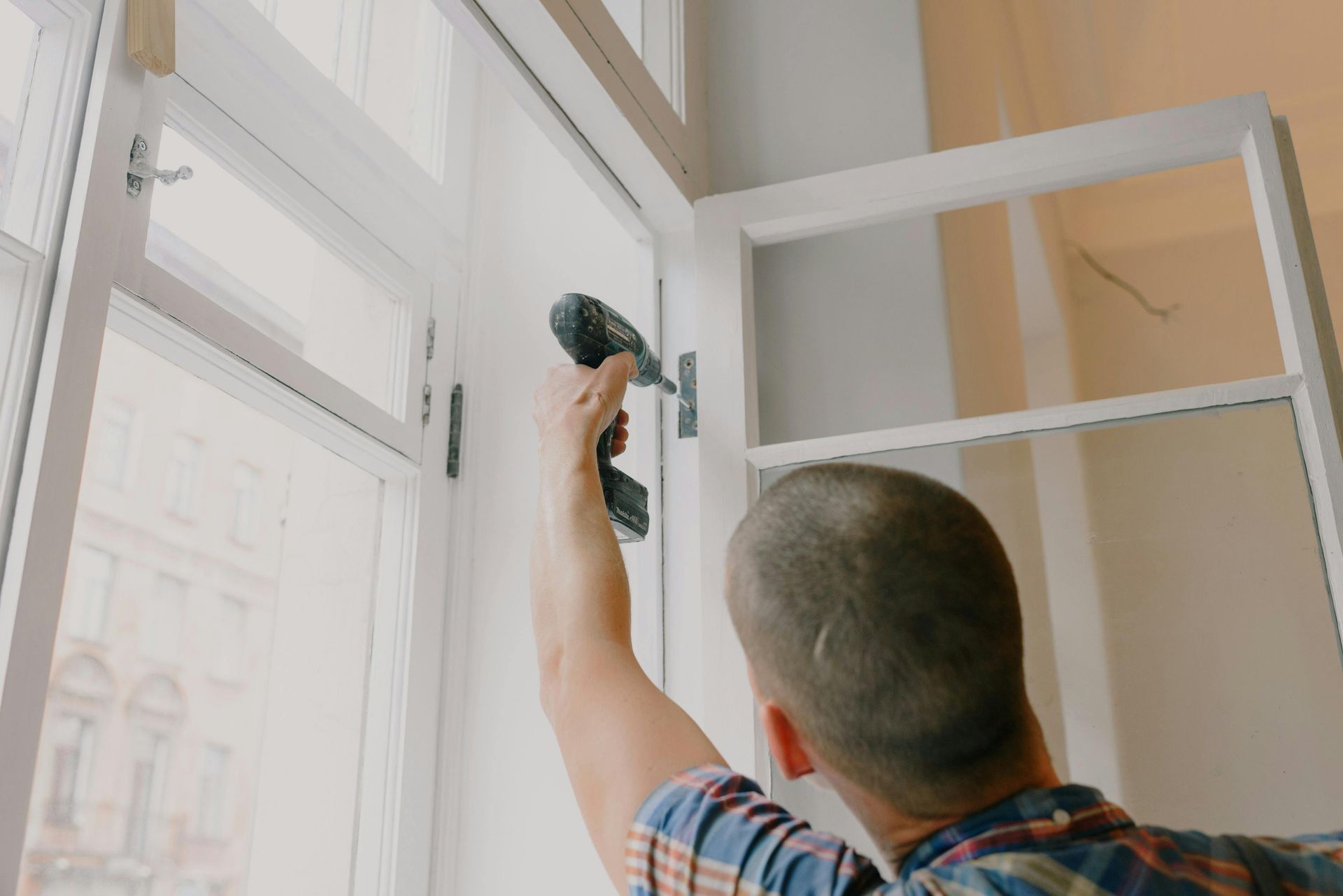 Person using a drill on a white window frame, presumably installing or repairing it. The setting appears to be indoors with natural light.