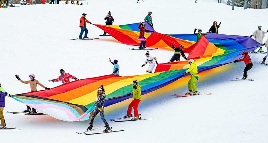 People on skis holding a large rainbow Pride flag on a snowy slope. Aspen Gay Ski Week