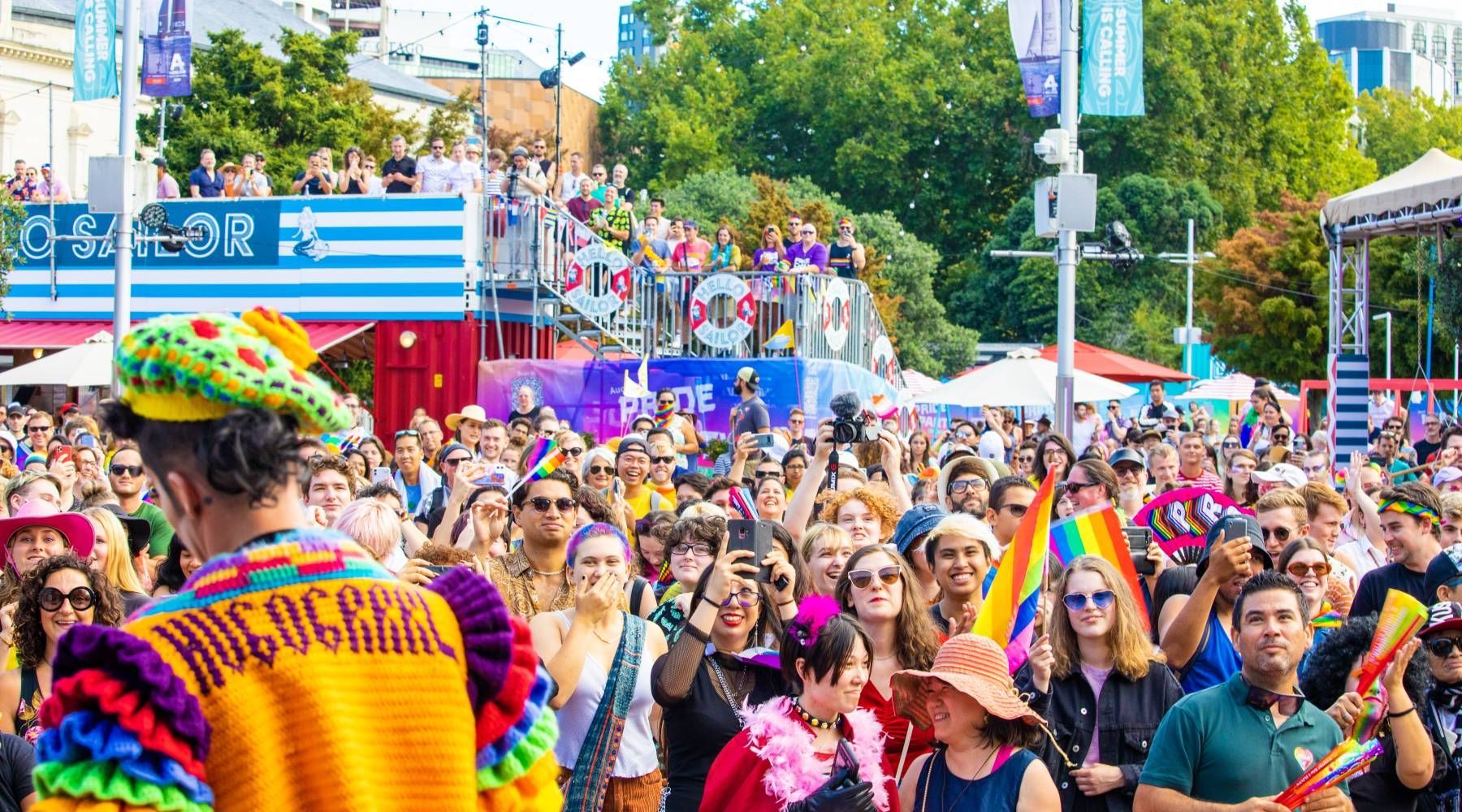 A crowd celebrating at a festival with a person in a colorful crochet jacket in the foreground. Auckland pride