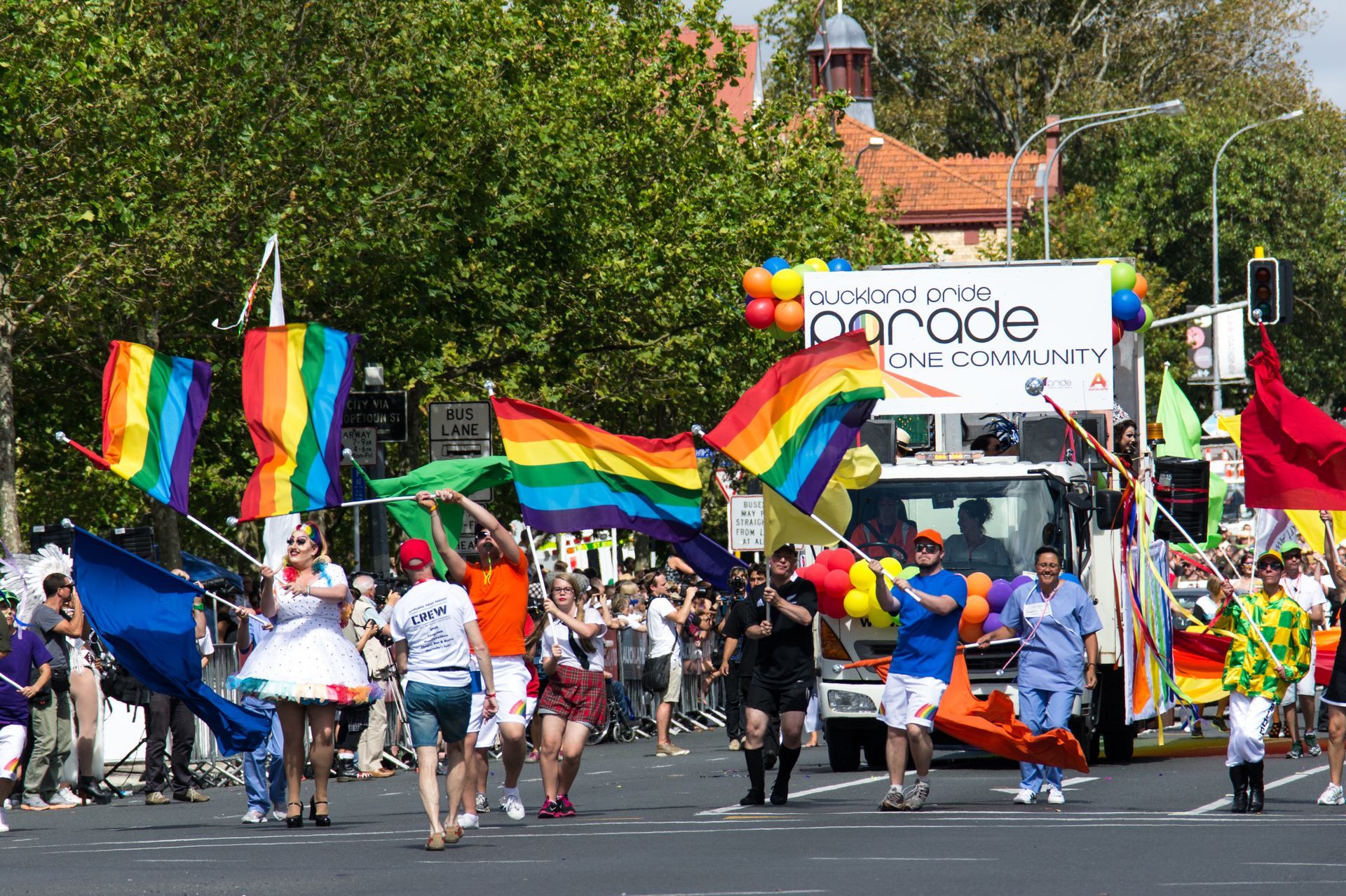 A parade featuring people walking down a street carrying rainbow flags, near a vehicle with 