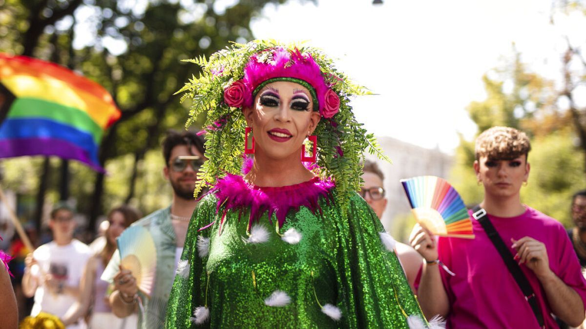 Drag queen in green sequined dress, pink accents, floral headpiece at a pride parade. Rainbow flag and people in background.