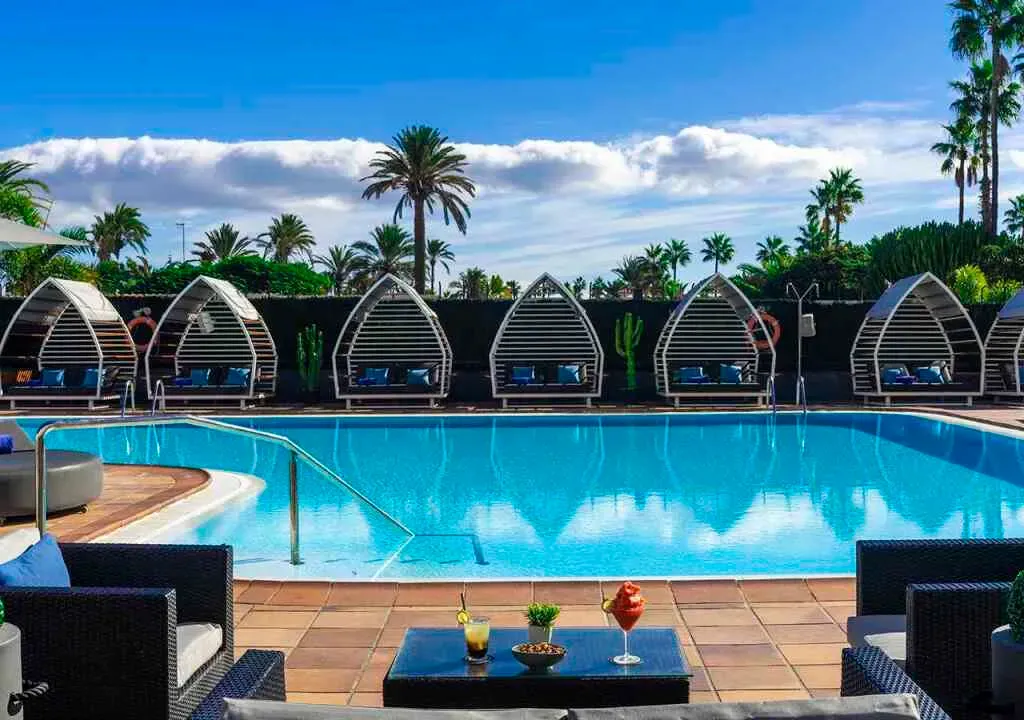 A bright swimming pool with cabanas lined along the back, viewed from a terrace with seating and drinks. Maspalomas
